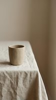 Minimalist ceramic cup in natural beige tone sitting on cream linen tablecloth against neutral wall background. Suitable for home decor, lifestyle, and product photography.