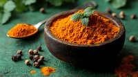 Ground turmeric powder in rustic clay bowl with mint leaves and scattered black peppercorns on dark green textured background, macro food photography.