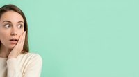 Surprised young woman with hand on face against mint green background, expressing shock and astonishment for advertising and marketing campaigns.