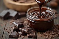 Melted chocolate pouring into glass jar surrounded by broken chocolate pieces and cocoa powder on rustic wooden surface for baking and confectionery.