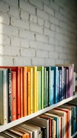 Colorful books arranged on white shelf against brick wall creating rainbow spectrum from red to purple for library or educational design.