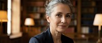 Confident mature Caucasian businesswoman with gray hair in elegant black blazer sitting in upscale office interior with warm lighting and brick walls.