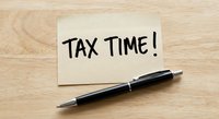 Handwritten tax time reminder note with black pen on wooden desk surface for accounting and financial planning preparation.