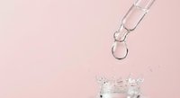 Glass dropper dispensing clear liquid into jar against soft pink background for skincare cosmetic product photography.