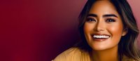 Young Asian woman with bright smile wearing yellow top against burgundy background, expressing joy and confidence in professional headshot style portrait.