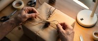 Close-up of hands wrapping kraft paper gift box with dried lavender sprigs and twine, workspace with tape and supplies visible, warm natural lighting.