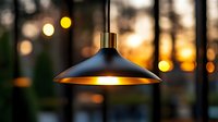 Modern black pendant lamp with gold interior illuminated against blurred sunset bokeh background, creating warm atmospheric lighting for home or restaurant decor.