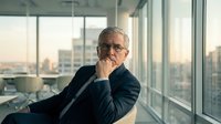 Mature male professional with gray hair and glasses wearing dark business suit, sitting by floor-to-ceiling windows in high-rise office overlooking cityscape, hand near face in thoughtful pose.