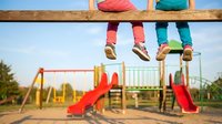 Two children sitting on wooden playground equipment wearing colorful clothing and shoes with playground structures in background on sunny day.