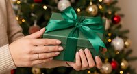 Hands holding wrapped gift box with green ribbon bow near decorated Christmas tree with warm lights creating festive holiday atmosphere for celebrations.