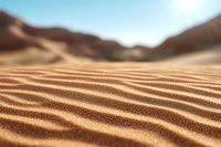 Close-up view of rippled sand dunes with soft waves and patterns in golden desert landscape against blurred mountainous background under blue sky.