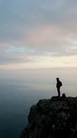 Silhouetted figure standing on rocky cliff edge overlooking calm ocean waters at dawn or dusk, conveying solitude, contemplation, and adventure. Suitable for travel, wellness, and motivational content.