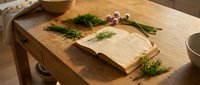 Wooden cutting board with fresh dill herbs and open recipe book on kitchen counter, cooking preparation and culinary planning concept.