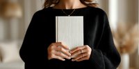 Woman in black sweater holding blank book with minimalist interior background, showcasing elegant jewelry and manicured nails.