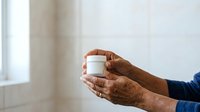 Middle-aged man with dark skin tone holding white cosmetic jar with both hands in bright interior space. Skincare product presentation for advertising and marketing purposes.