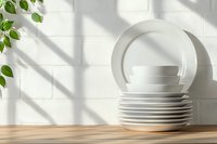Stack of clean white ceramic plates and cup on wooden table with natural sunlight casting shadows through window, green plant leaves add fresh accent.