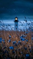 Lighthouse glowing at dusk with illuminated tower behind wild grass and blue flowers under starry night sky, moody coastal landscape atmosphere.