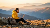 Young woman in yellow jacket working on laptop computer while sitting on mountain peak during golden hour sunset with scenic valley landscape background.