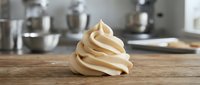 Creamy swirl of whipped cream or frosting on wooden surface with kitchen equipment blurred in background, suitable for baking and culinary content.