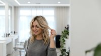 Young blonde Caucasian woman touching her hair in modern beauty salon interior with professional styling chairs and bright lighting for hair care services.