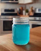 Glass mason jar filled with vibrant turquoise liquid on wooden kitchen counter, blurred modern appliances in background create depth and atmosphere.
