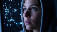 Young Caucasian woman with hood gazing at futuristic holographic digital interface with glowing blue data particles and network connections in dark environment.