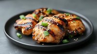 Grilled teriyaki chicken thighs garnished with sesame seeds and green onions served on black plate, close up view of juicy glazed meat with charred edges.