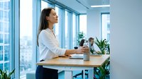 Professional businesswoman standing at modern office desk looking out window with natural light and urban view in contemporary workplace.