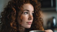 Young Caucasian woman with natural curly red hair and freckles gazing thoughtfully in soft kitchen lighting, creating intimate portrait atmosphere.