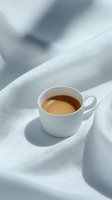 White cup of creamy coffee on soft white fabric with gentle shadows creating minimalist composition for lifestyle and food blogs.