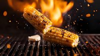 Grilled corn on cob with butter falling in motion against flaming grill background, bright orange sparks flying, dramatic dark food photography with fire elements.