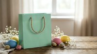 Mint green gift bag with rope handles surrounded by colorful Easter eggs and white flowers on wooden table near bright window for spring celebration.