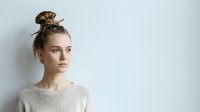 Young woman with light brown hair styled in a top knot bun, fair complexion, wearing a cream knit sweater, looking thoughtfully to the side against a white background.