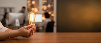 Hand holding glowing vintage Edison bulb on wooden table with blurred office background, symbolizing innovation, creativity, and bright business ideas.