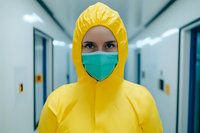 Healthcare worker in yellow protective hazmat suit and green surgical mask standing in hospital corridor with fluorescent lighting and blue walls.