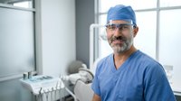 Middle-aged male dentist of South Asian ethnicity wearing blue surgical scrubs, head cap and protective eyeglasses, smiling confidently in modern dental clinic with equipment and window background.