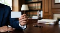 Businessman in navy suit holding blank white business card at wooden desk with office background and bookshelf, corporate identity and professional branding concept.
