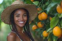 Young African American woman in straw hat smiling while standing near orange tree in garden, fresh citrus fruits and green leaves background, summer harvest concept.