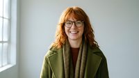 Smiling middle-aged Caucasian woman with red curly hair and glasses wearing green coat indoors by window for professional business portrait.