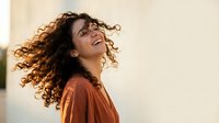 Young Hispanic woman with curly hair laughing joyfully outdoors, golden sunlight, carefree emotion, copy space on light background.