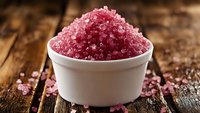 Pink Himalayan salt crystals in white ceramic bowl on rustic wooden table, macro view with scattered grains showing texture and mineral-rich coloring.