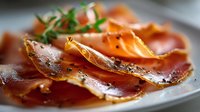 Thinly sliced cured ham with herbs on white plate, close-up view showing delicate texture and rich color of premium charcuterie.