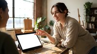 Young woman with dark hair and South Asian ethnicity, wearing a beige cardigan, smiling while gesturing during a video call on laptop in a modern home office with plants and natural light.