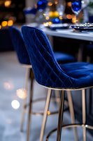 Luxurious navy blue velvet bar stool with quilted diamond pattern and gold metal legs against blurred bokeh restaurant background, elegant dining atmosphere.