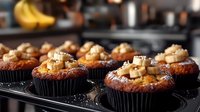Chocolate banana cupcakes topped with caramel pieces and nuts in black liners, fresh baked desserts on dark baking tray with blurred kitchen background.