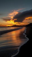 Dramatic golden sunset over coastal beach with dark storm clouds and reflective wet sand creating mirror effect as waves curve along shoreline during magic hour.