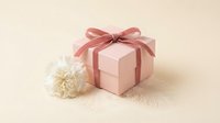 Pink gift box with coral ribbon bow and white carnation flower on neutral beige background for special occasions and celebrations.