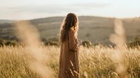 Young woman with long brown hair in flowing dress standing in golden wheat field during sunset with warm sunlight and rolling hills in background.