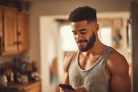 Smiling mixed-race man in tank top using smartphone in modern kitchen. Happy young adult male texting on mobile phone at home during morning routine.