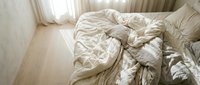 Unmade bed with crumpled beige bedding and pillows in bright morning light from window, conveying comfort and relaxation in bedroom interior design.